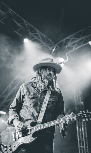 Man with long hair and a hat playing electric guitar with a pick on stage