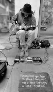 Man busking using a chain of guitar pedals and a capo